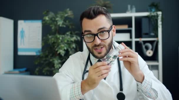 Ritratto di giovane medico amichevole che mostra pillole e parla guardando la macchina fotografica in clinica — Video Stock