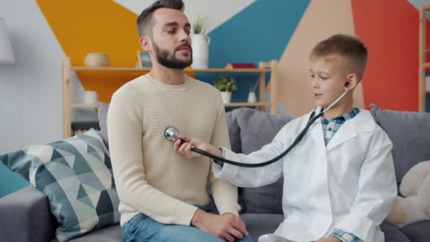 Far och son leker läkare och patient medan pojken använder stetoskop sedan skrattar hemma — Stockvideo