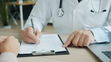 Sağlık raporunu dolduran erkek doktorun yakın plan resmi. Ofisteki kadın hastayla konuşuyor.