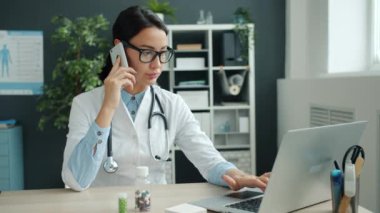 Genç bayan doktor cep telefonuyla konuşuyor ve ofiste çalışırken laptop kullanıyor.