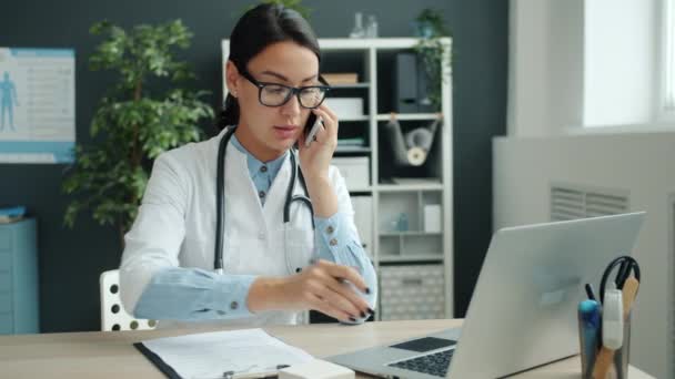 Chica seria en vestido blanco médico hablando en el teléfono móvil y el uso de la computadora portátil — Vídeo de stock