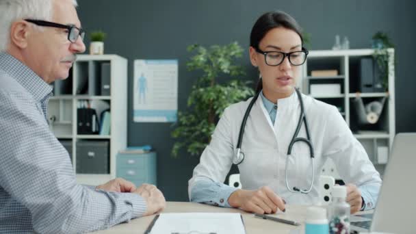 Καλός γιατρός μιλάει σε παλιό ασθενή δείχνει οθόνη laptop στη συνέχεια κρατώντας το χέρι στο γραφείο — Αρχείο Βίντεο