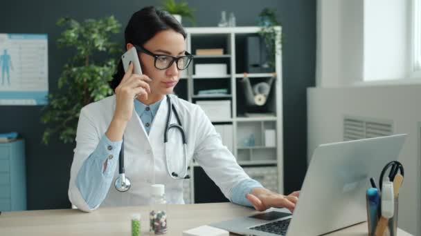 Genç bayan doktor cep telefonuyla konuşuyor ve ofiste çalışırken laptop kullanıyor. — Stok video