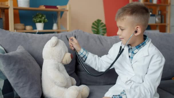 Divertido niño jugando con osito de peluche examinando juguete con estetoscopio disfrutando del juego solo — Vídeo de stock