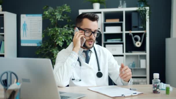 Młody, pogodny lekarz rozmawiający przez telefon komórkowy, piszący na wykresie i korzystający z laptopa w biurze — Wideo stockowe