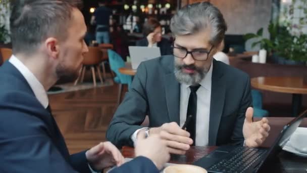 Los hombres de negocios serios hablando y haciendo gestos con el ordenador portátil en la reunión de negocios en la cafetería — Vídeos de Stock