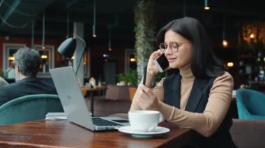 Neşeli genç bayan cep telefonuyla konuşuyor ve kafede dizüstü bilgisayarla çalışıyor.