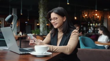 Neşeli bayan girişimci kafeteryada gülümseyen ekranlara dokunarak akıllı telefon kullanıyor