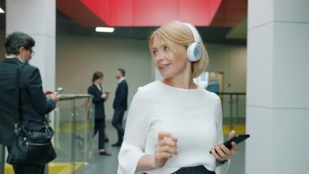 Attractive mature businesswoman in headphones walking and dancing in lobby holding smartphone — Stock Video