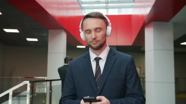 Доллі знімок молодого бізнесмена, який слухає музику в навушниках і використовує смартфон у вестибюлі — стокове відео