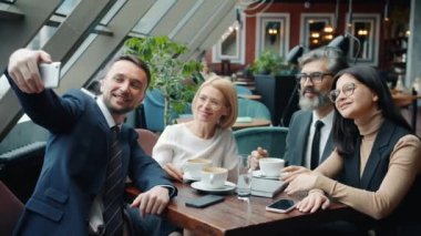 Genç adam bir grup iş ortağıyla restoranda cep kamerası kullanarak selfie çekiyor.