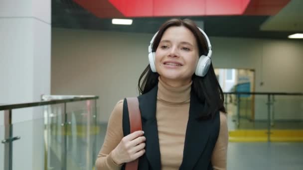 Joven dama de negocios alegre caminando en el vestíbulo con auriculares usando teléfono inteligente — Vídeo de stock