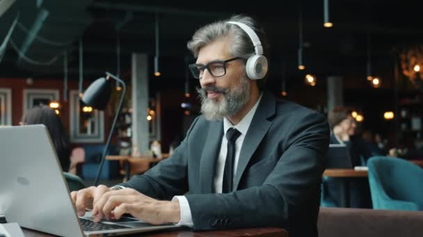 Man i kostym arbetar med bärbar dator och njuter av musik i hörlurar på café — Stockvideo