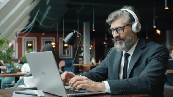 Uomo d'affari maturo con le cuffie che lavorano con il computer portatile nel ristorante — Video Stock