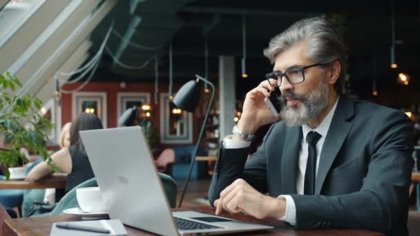 Uomo d'affari allegro che parla sul telefono cellulare e utilizza il computer portatile nel ristorante — Video Stock