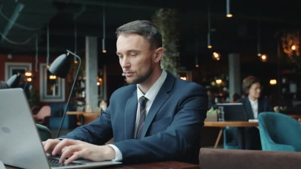 Joven serio en traje usando el ordenador portátil escribiendo trabajando solo en el restaurante — Vídeo de stock