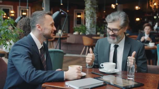 Hombres de negocios felices hablando mostrando los pulgares hacia arriba y tomando café en la cafetería — Vídeos de Stock