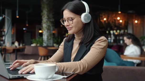 Счастливая деловая леди, использующая ноутбук в кафе и слушающая музыку через беспроводные наушники — стоковое видео