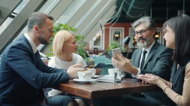 Bir grup neşeli kadın ve erkek akıllı telefon kullanıyor ve kafede sohbet ediyorlar.