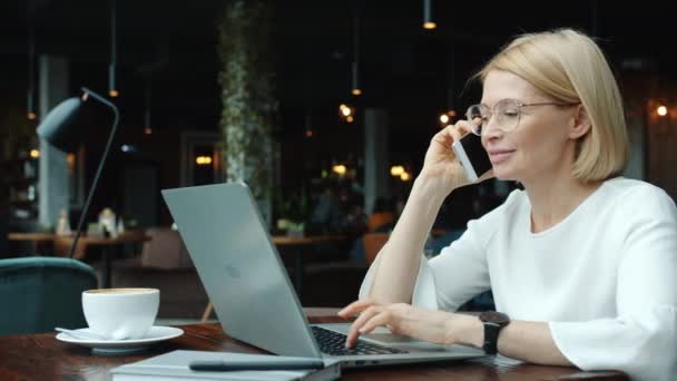 Administrador alegre hablando en el teléfono móvil y el uso de la computadora portátil en la cafetería — Vídeo de stock
