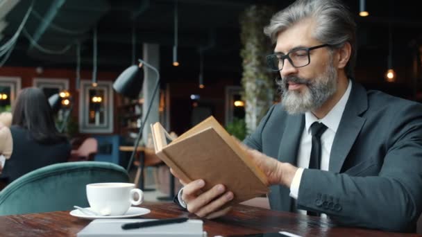 Mature man in glasses and suit reading book at table in restaurant focused on literature — Stock Video