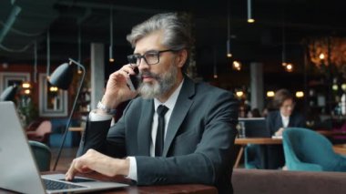 Girişimci olgun sakallı adam cep telefonuyla konuşuyor ve kafede dizüstü bilgisayarla çalışıyor.