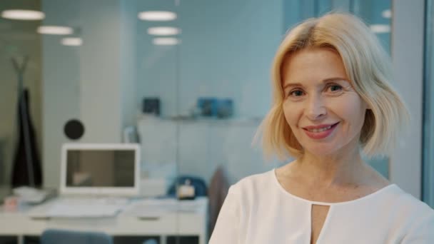 Hidasliikkeinen muotokuva kypsä blondi työntekijä hymyilee lasiseinätoimistossa — kuvapankkivideo