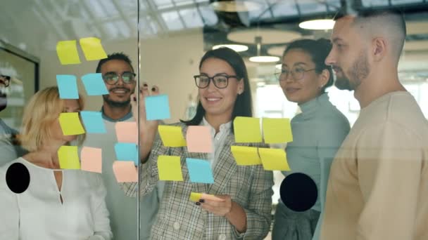 Empresaria pegando notas en el tablero de cristal discutiendo ideas con colegas aplaudiendo de la mano — Vídeo de stock