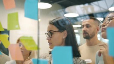 Takım lideri cam tahtada yapışkan notlarla çalışıyor ve ofisteki iş arkadaşlarıyla konuşuyor.