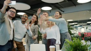 Mutlu iş arkadaşları iş yerindeki akıllı telefonu kullanarak online video görüşmesi yapıyor ve gülüp el sallıyorlar.