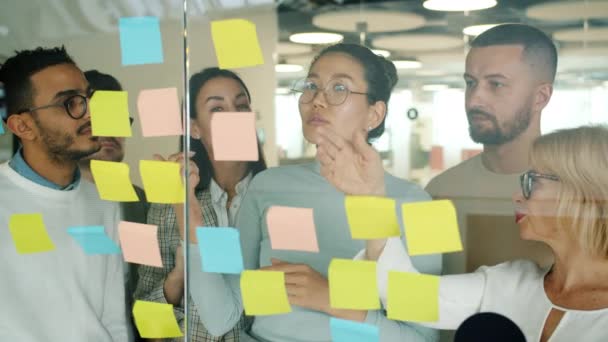 Jóvenes creativos hombres y mujeres que trabajan con notas adhesivas en el tablero de cristal discutiendo ideas — Vídeo de stock