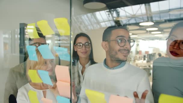 Arab man talking to colleagues discussing ideas on sticky notes working with glassboard — Stock Video