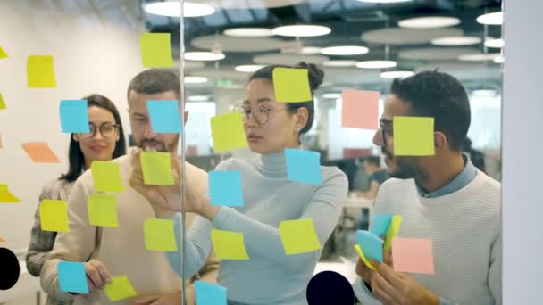Творческая команда бизнесменов приклеивает заметки на стеклянную доску для мозгового штурма в офисе — стоковое видео
