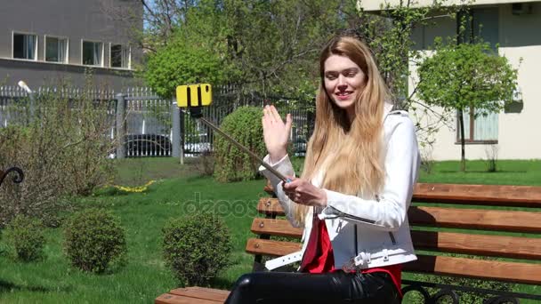 Video uitzending. Jonge blonde vrouw communiceert op mobiel zittend op bankje in park — Stockvideo