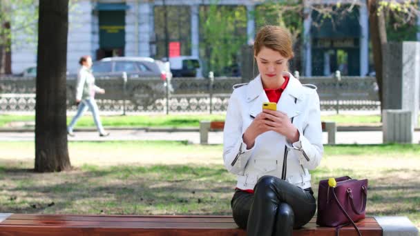 Videokonferenz. junges schönes blondes Model kommuniziert auf Handy sitzend auf Bank im Park — Stockvideo