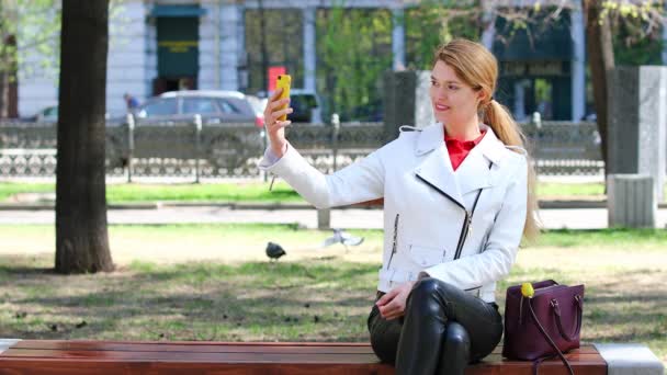 Videokonferenz. junges schönes Mädchen kommuniziert auf Handy sitzend auf Bank im Park — Stockvideo