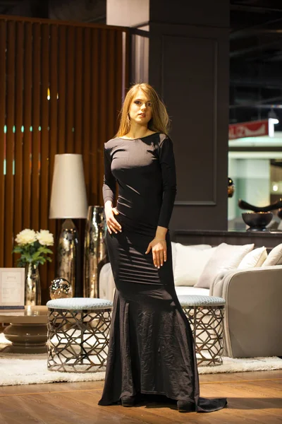 Sexy blonde woman in a black evening dress posing in a furniture — Stock Photo, Image