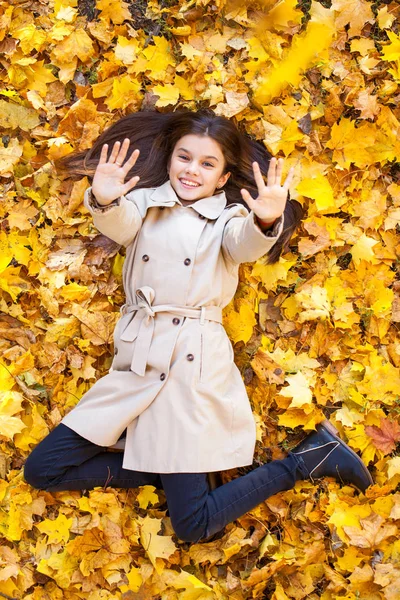 Mladá krásná holčička v béžovém kabátě ležící na žlutých listech — Stock fotografie