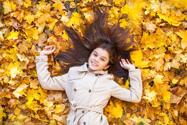 Giovane bella bambina in cappotto beige sdraiata su foglie gialle — Foto Stock