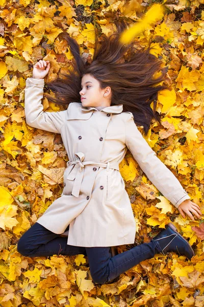 Giovane bella bambina in cappotto beige sdraiata su foglie gialle — Foto Stock