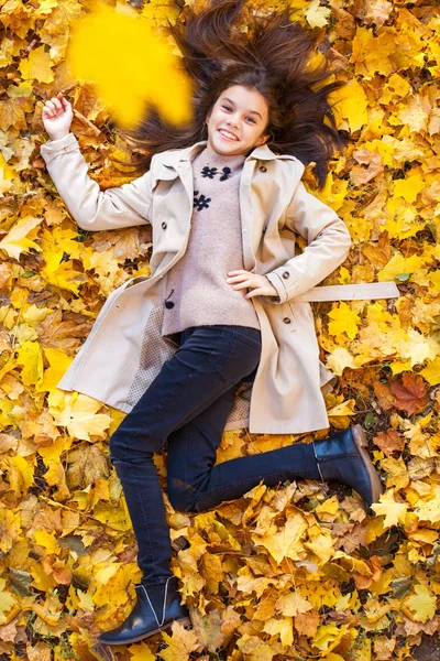 Young beautiful little girl in beige coat lying on yellow leaves — Stockfoto