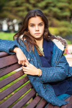 Mavi ceketli küçük kız bir bankta oturuyor.