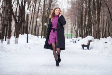 Uzun siyah ceketli, kış parkında poz veren şık bir genç kızın tam boy portresi.