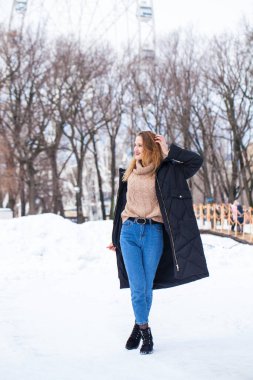 Uzun siyah ceketli, kış parkında poz veren şık bir genç kızın tam boy portresi.