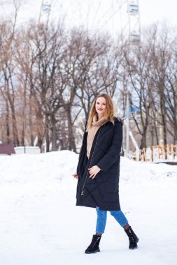 Uzun siyah ceketli, kış parkında poz veren şık bir genç kızın tam boy portresi.