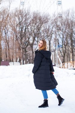 Uzun siyah ceketli, kış parkında poz veren şık bir genç kızın tam boy portresi.