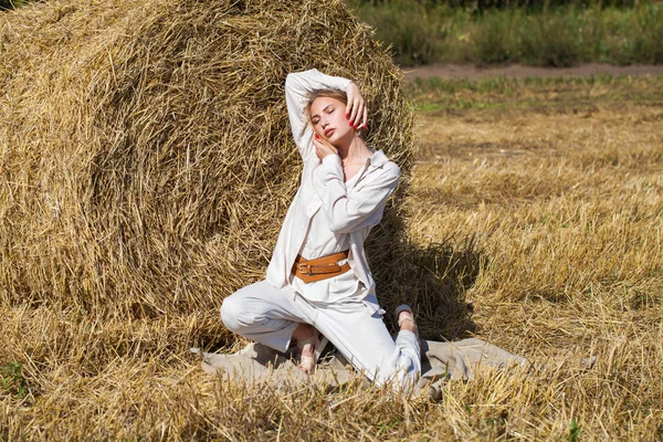 穿着米色西服的漂亮金发姑娘站在割破的田野里的干草堆后面 — 图库照片