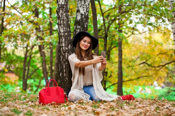 在秋天的森林里 戴着帽子的年轻漂亮的女人坐在手机上拍照 — 图库照片
