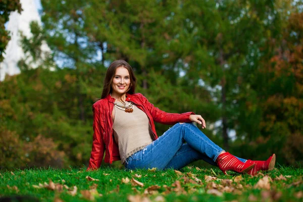 若いです美しいです女性で赤い革のジャケットと青いジーンズで秋の公園に座って — ストック写真