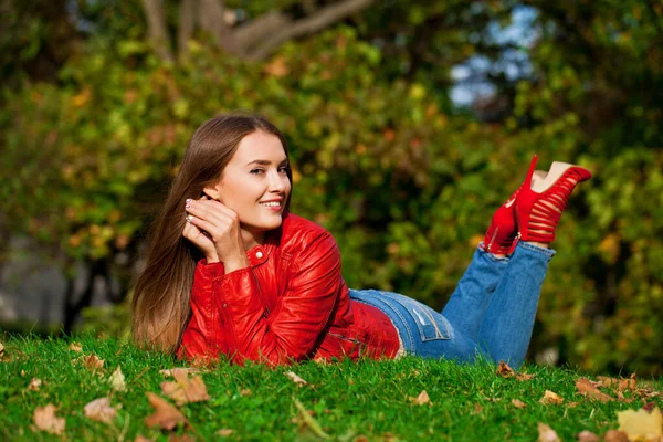 若いです美しいです女性で赤い革のジャケットと青いジーンズで秋の公園に座って — ストック写真